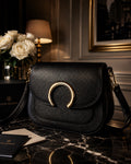 “Black leather Luxe Maison crossbody bag with gold horseshoe buckle on marble table beside white roses”