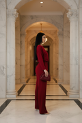 A woman in a long-sleeve, wide-leg burgundy jumpsuit stands in profile in a grand marble hallway with arches and columns, holding a matching burgundy clutch.