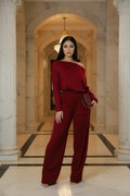 A woman stands in a grand marble hallway wearing a deep burgundy one-shoulder jumpsuit with wide-leg pants and holding a matching clutch bag.