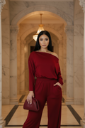 A woman stands in a grand marble hallway wearing a deep burgundy off-the-shoulder jumpsuit and holding a matching burgundy clutch.