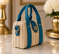 A small woven straw handbag with teal blue fabric handles and trim, displayed on a polished marble surface with a gold lamp and white flowers in the background.