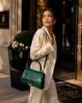 A woman in a cream-colored blazer and trousers carries a structured emerald green crocodile-embossed leather handbag with gold-tone hardware, walking in front of a luxury building entrance.