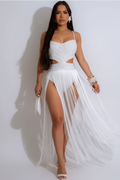 A woman modeling a white two-piece outfit consisting of a cropped camisole top with cutouts and a matching high-slit maxi skirt with a flowing tulle overlay, set against a plain light gray studio background.