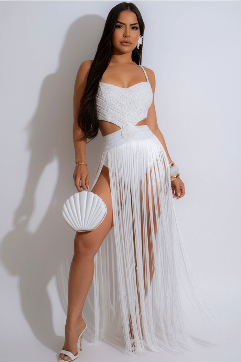 A woman modeling a white two-piece outfit consisting of a textured crop top and a high-slit maxi skirt with long fringe, holding a matching white shell-shaped handbag against a plain white studio background.