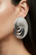 A close-up of a model's ear wearing a large, sculptural spiral earring. The earring features a polished silver-tone metal spiral structure with a textured, diamond-paved outer surface.
