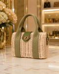 A small woven straw handbag with olive green fabric handles and side panels, featuring a gold-tone snap button closure, displayed on a marble countertop in a boutique setting.