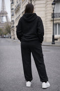 Person wearing a black hoodie and pants standing on a street with a blurred Eiffel Tower in the background