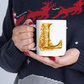 Person holding a mug with a decorative letter 'L' design, wearing a dark sweater with red deer pattern.