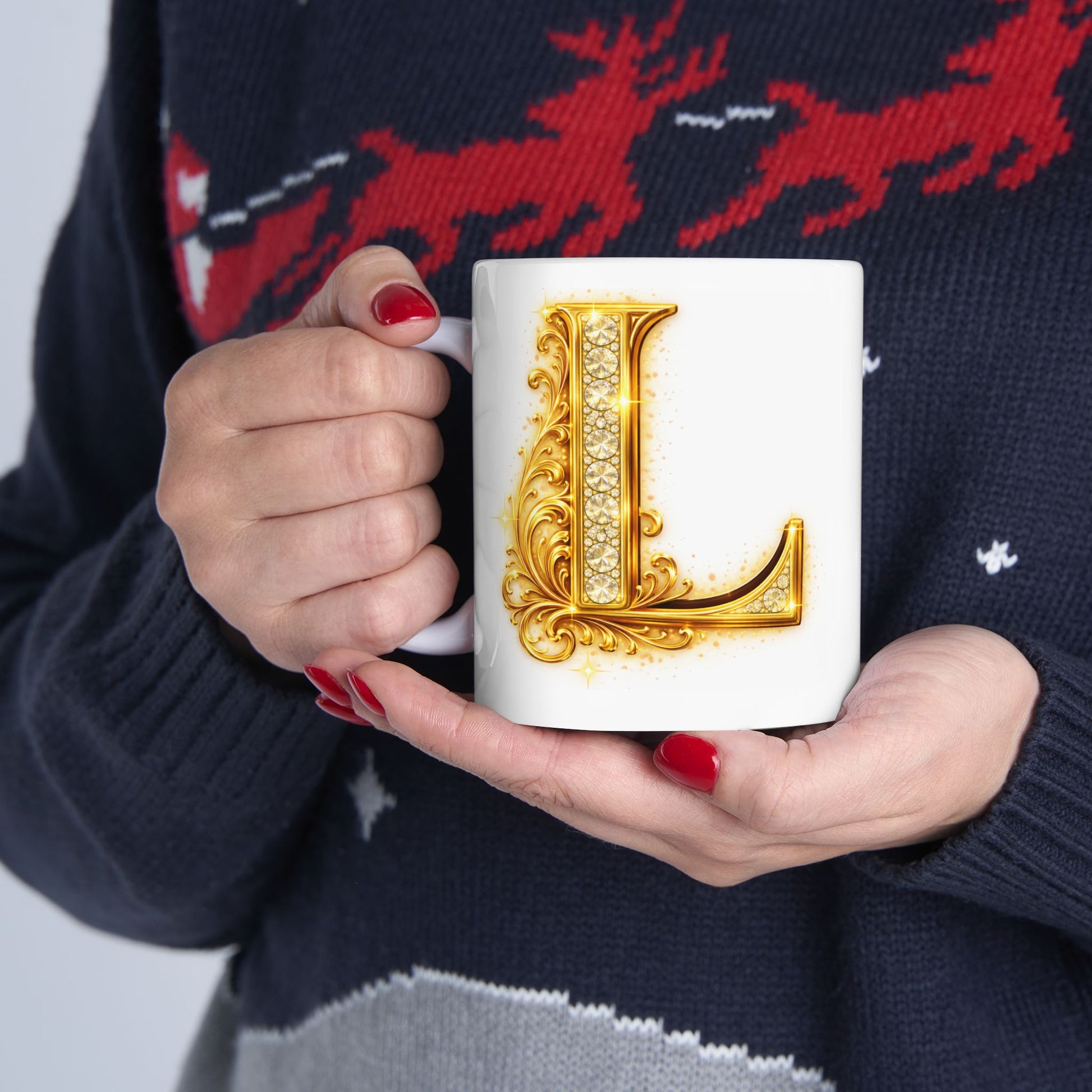 Person holding a mug with a decorative letter 'L' design, wearing a dark sweater with red deer pattern.