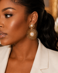 A close-up of a woman wearing a pair of gold sunburst dangle earrings with white pearl centers, paired with a white blazer.