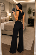 A woman takes a mirror selfie in a bedroom, wearing a black sleeveless crop top and matching high-waisted wide-leg pants.