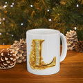 White mug with gold letter 'L' design in front of a Christmas tree with pinecones