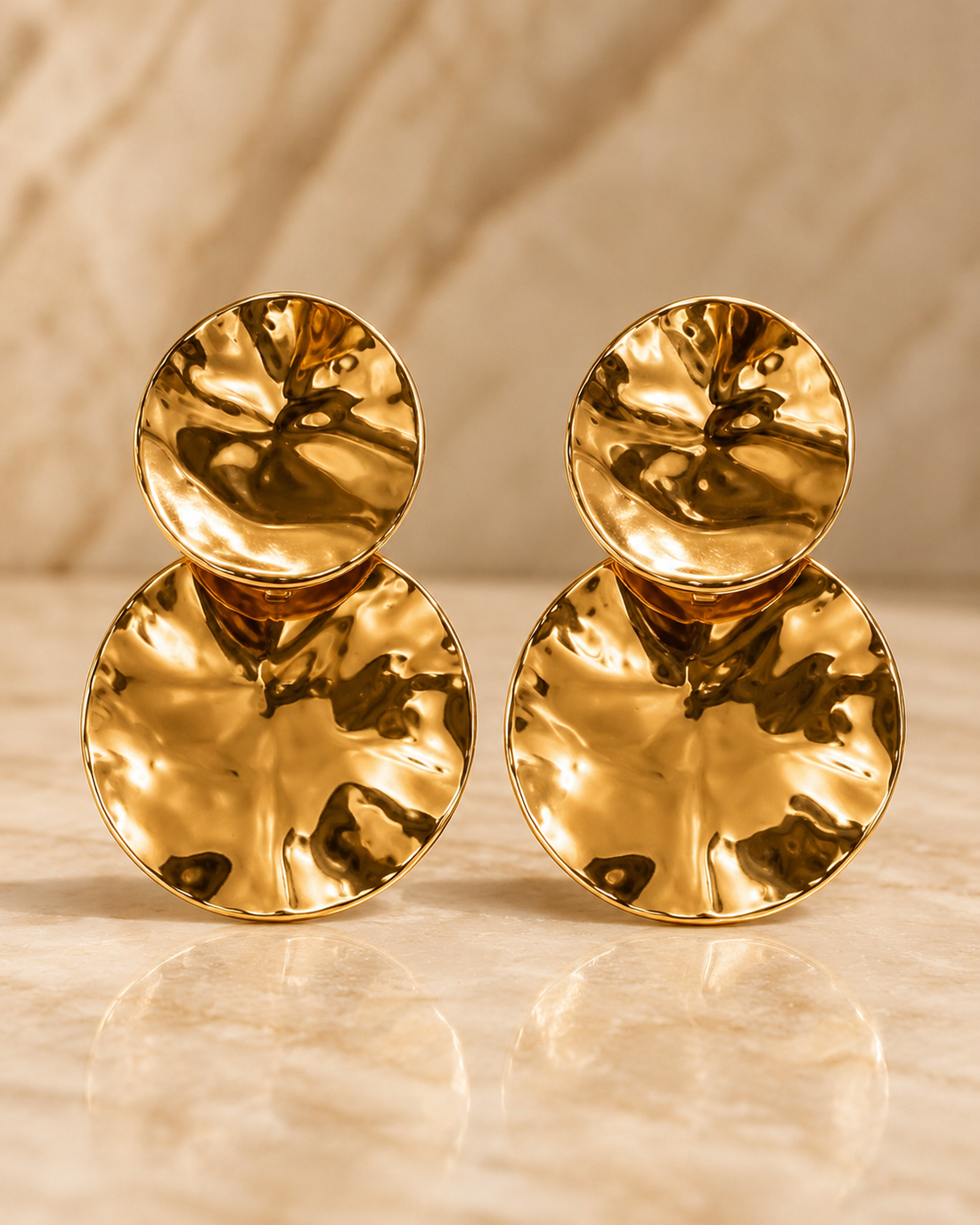 A close-up portrait of a woman wearing large, chunky gold disc earrings with a faceted, textured surface. She is dressed in a champagne-colored one-shoulder gown, set against a warm, golden background with soft bokeh lights.