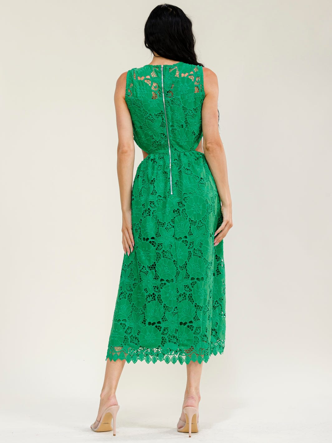 A woman walking on a city sidewalk wearing a sleeveless bright green lace midi dress with a fitted bodice and flared skirt. She is carrying a small woven beige handbag and wearing beige strappy heels.