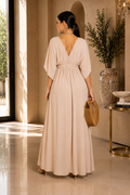 A woman stands with her back to the camera wearing a floor-length beige dress with a deep V-neckline, short draped sleeves, and a gathered waist. She is posed in a sunlit interior with a woven clutch and heels.