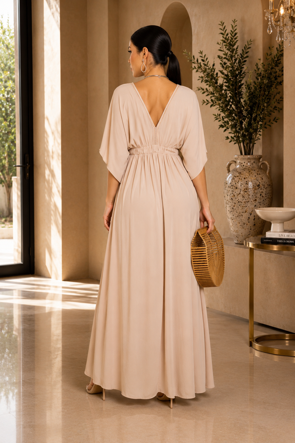 A woman stands with her back to the camera wearing a floor-length beige dress with a deep V-neckline, short draped sleeves, and a gathered waist. She is posed in a sunlit interior with a woven clutch and heels.