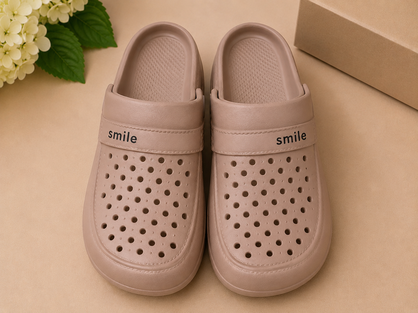 Pair of beige clogs with 'smile' text on a beige surface with flowers and a box in the background.