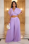 A woman models a lavender two-piece matching set consisting of a short-sleeve wrap-style crop top and high-waisted wide-leg pants, standing between white columns in a grand interior hallway.
