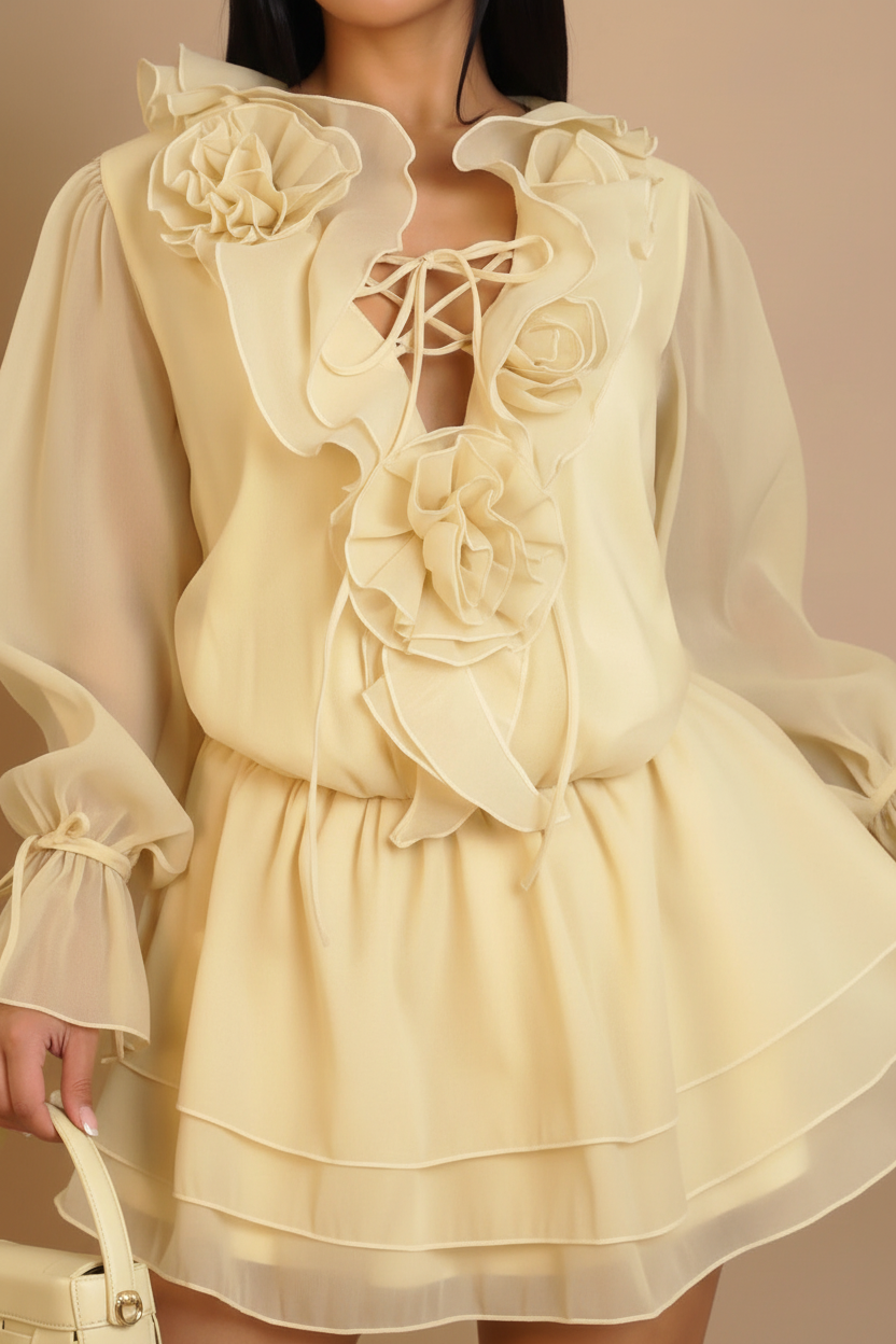 A close-up of a model wearing a cream-colored mini dress with large 3D floral appliqués, ruffled collar, lace-up neckline, and a tiered skirt.