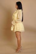 A woman in profile wearing a pale yellow ruffled long-sleeve mini dress with a tiered skirt, holding a matching cream handbag, against a solid beige studio background.