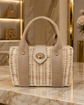 A structured wicker handbag with woven beige handles and a beige front flap, featuring gold-tone hardware, displayed on a marble countertop in a luxury interior setting.