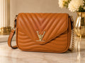A tan quilted leather shoulder bag with a chevron pattern, gold-tone LV logo clasp, and a gold chain strap, displayed on a marble surface with a blurred background.