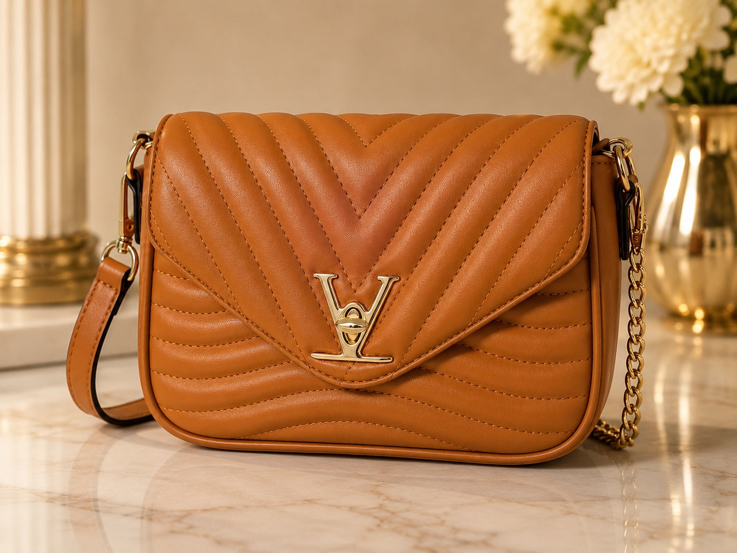 A tan quilted leather shoulder bag with a chevron pattern, gold-tone LV logo clasp, and a gold chain strap, displayed on a marble surface with a blurred background.