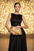 A woman wearing a sleeveless black satin gown holds a textured gold clutch bag against a warm, glowing bokeh background.