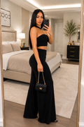A woman takes a mirror selfie in a modern bedroom, wearing a black two-piece outfit consisting of a strapless crop top and matching wide-leg pants. She is holding a small black quilted handbag with a gold chain strap.