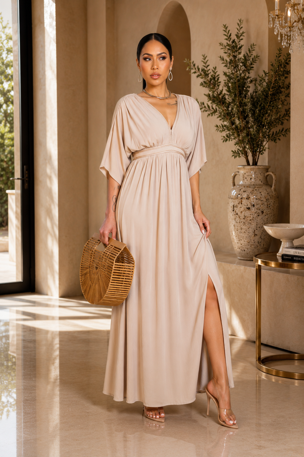 A woman models a floor-length beige maxi dress with a V-neckline, short flutter sleeves, a matching waist tie, and a high side slit. She is holding a round woven straw clutch and posing in a warm, elegant interior setting.