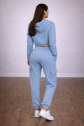 Woman wearing a light blue tracksuit standing in a room with wooden floor and white walls.

