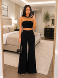 A woman takes a mirror selfie in a modern bedroom, wearing a black two-piece outfit consisting of a strapless bandeau crop top and matching high-waisted wide-leg pants. She is accessorized with a gold choker necklace, a small black quilted handbag with a gold chain strap, and black high-heeled sandals.