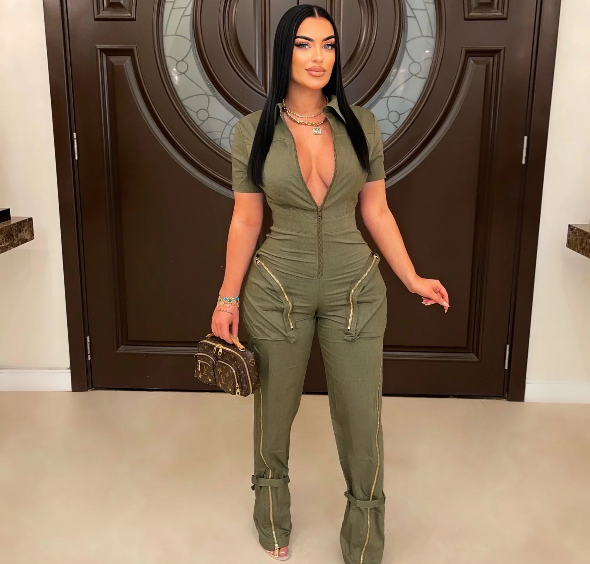 Woman in a green jumpsuit holding a handbag in front of a decorative door.