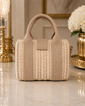 A beige woven straw handbag with a top handle and a matching leather strap detail, displayed on a polished marble surface with elegant gold-toned decor in the background.
