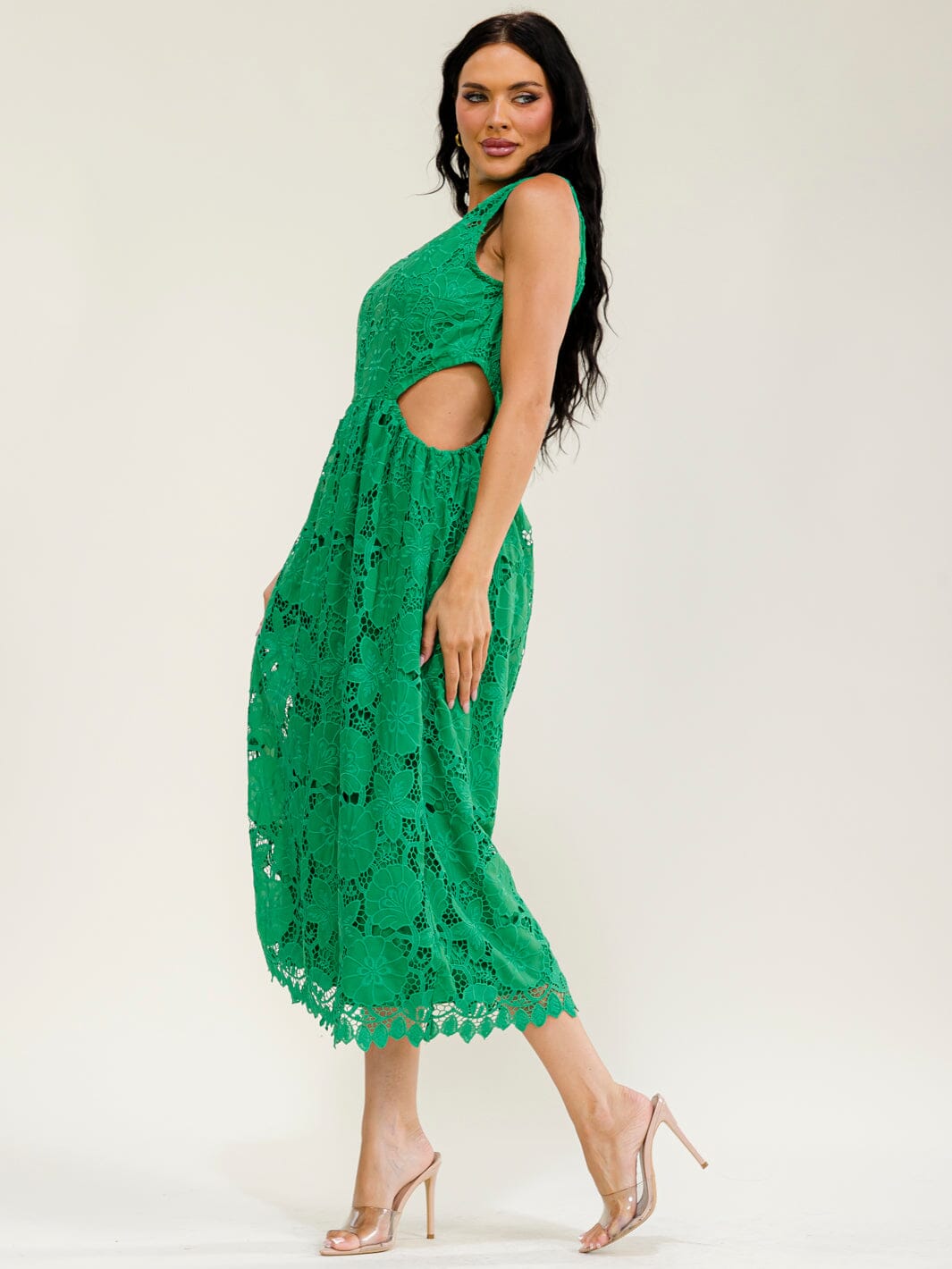 A woman modeling a bright green sleeveless midi dress with a cutout at the waist and floral lace pattern, paired with clear high-heeled sandals against a plain light background.