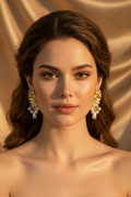 A close-up portrait of a woman with styled brown hair wearing ornate gold and pearl drop earrings against a warm beige satin fabric background.