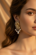 A close-up of a woman wearing an ornate gold and pearl traditional Indian earring, set against a warm, draped beige background.