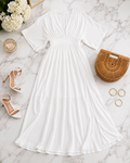 A white short-sleeve V-neck maxi dress with a smocked waist and pleated skirt, styled in a flat lay on a white marble background with white high-heeled sandals, a brown woven clutch, gold hoop earrings, and white flowers.