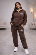 Woman wearing a brown tracksuit with 'alo' branding in a modern indoor setting.