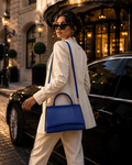 A woman in a cream-colored blazer and trousers walks past a black luxury car on a cobblestone street, carrying a structured royal blue leather shoulder bag with gold-tone hardware.