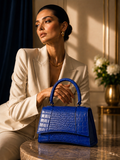 A woman in a cream blazer sits at a marble table holding a vibrant blue crocodile-embossed handbag with a top handle in a warm, elegant interior setting.