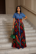 A woman stands on marble stairs wearing a long dress with a blue short-sleeve shirt bodice and a colorful geometric patterned skirt in red, green, blue, yellow, and black. She is holding a green handbag.
