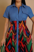 A woman wearing a short-sleeve button-front dress with a solid blue upper section and a colorful geometric patterned lower skirt in red, green, black, orange, and beige, against a plain beige background.