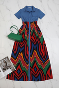 A short-sleeve collared dress laid flat on a white marble surface, featuring a solid blue upper bodice and a vibrant multicolor zigzag patterned skirt in red, blue, green, orange, and black. A green handbag and a magazine are styled alongside the dress.