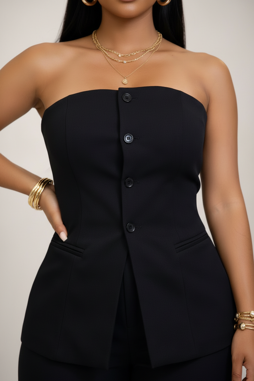 A close-up of a model wearing a black strapless tube top with a button-front detail and structured shoulders, styled with gold jewelry against a neutral beige background.