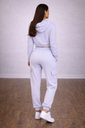 Woman wearing a light gray tracksuit standing in a room with wooden floor and white walls.