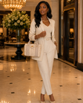 A woman stands in a luxury hotel lobby wearing a matching cream-colored three-piece suit, including a blazer, vest, and trousers, paired with a white top and beige heels. She is holding a structured cream handbag with gold hardware.