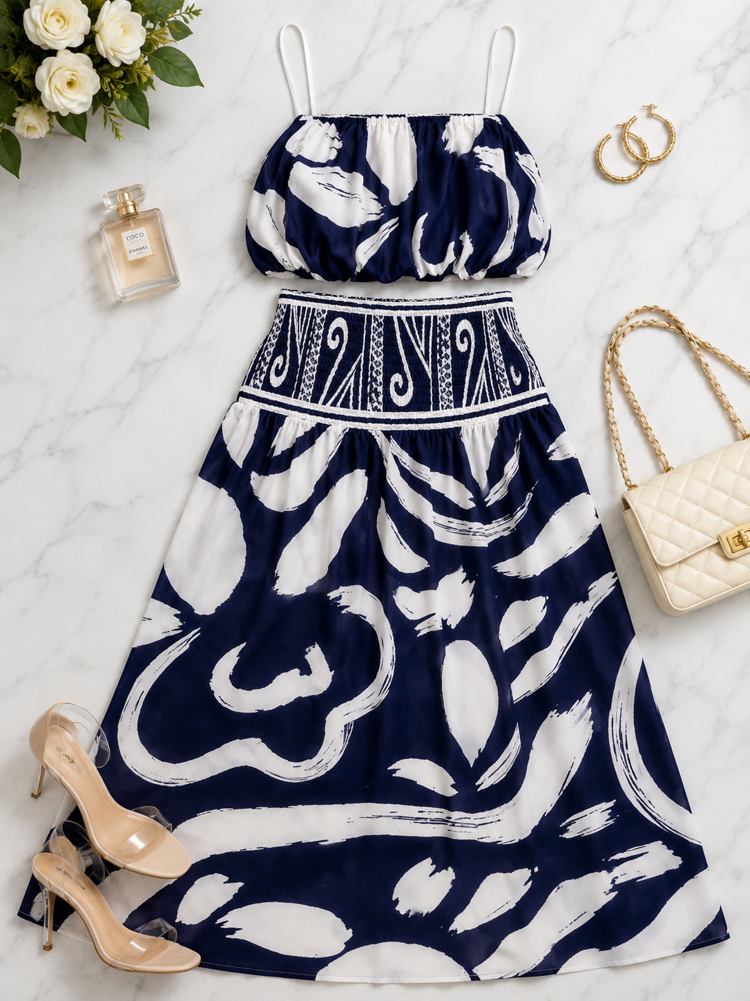 A navy blue and white abstract print two-piece set consisting of a sleeveless crop top with spaghetti straps and a matching A-line skirt, styled in a flat lay with beige heels, a quilted cream handbag, gold hoop earrings, a perfume bottle, and white roses on a white marble background.