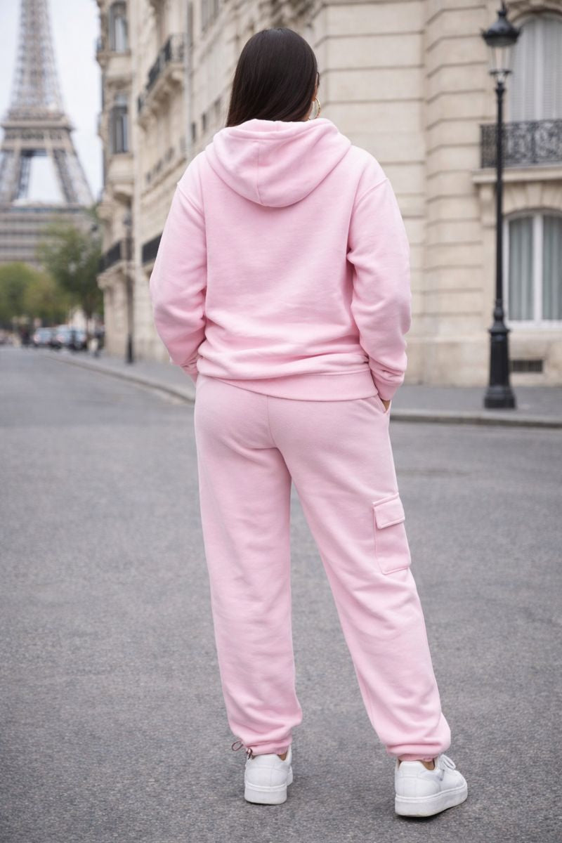 Person wearing a pink tracksuit standing on a street with a building and lamp post in the background.