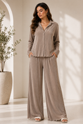 Woman wearing a beige button-up shirt and wide-leg pants in a bright room with a plant.